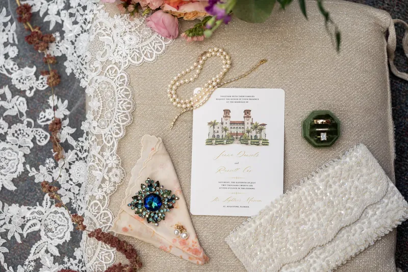 Flat lay of wedding details including lace, pearl necklace, sapphire ring, program, and flowers on neutral fabric.