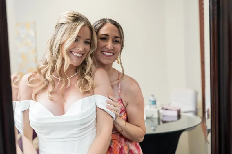 Jenna and her bridesmaid laugh together while getting ready, both dressed in white and coral.