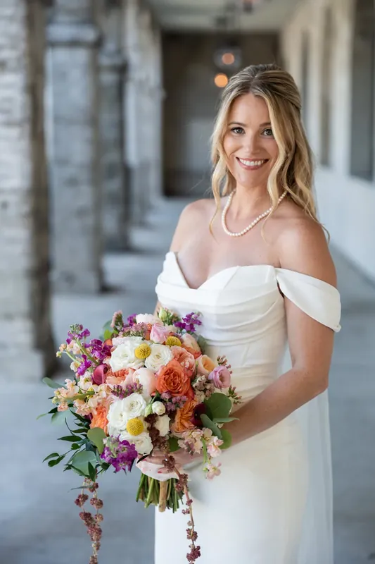 Jenna holds a vibrant bouquet of coral, pink, and cream flowers while wearing an off-shoulder white gown at the Lightner