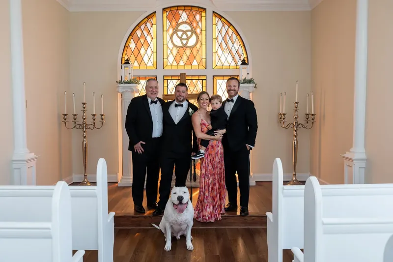 Jenna and Russell stand with their dog and wedding party in front of the chapel's stained-glass window at the museum.