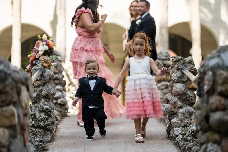 Young children walk down the covered courtyard passageway, the girl in a pink dress and boy in a tuxedo, flanked by family