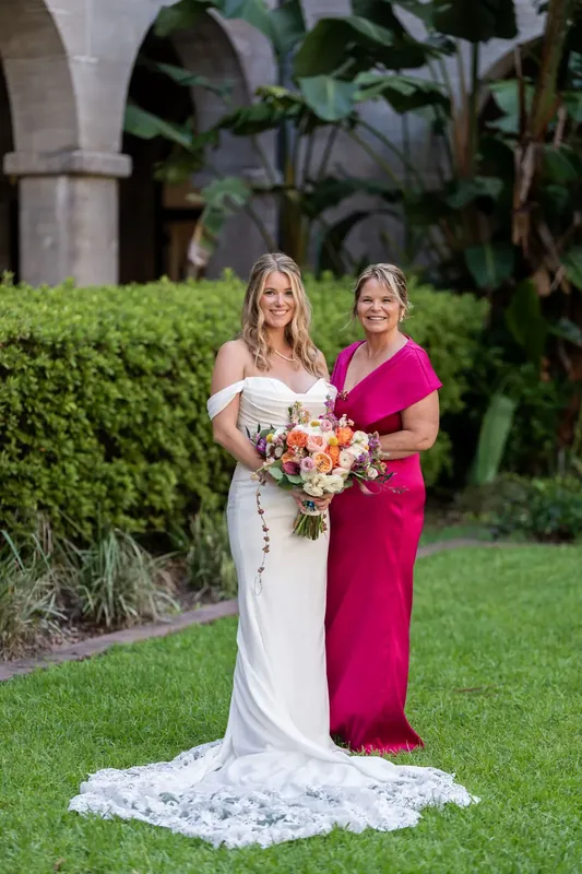 Jenna in her wedding dress stands beside a woman in a vibrant magenta gown, both holding the bridal bouquet.