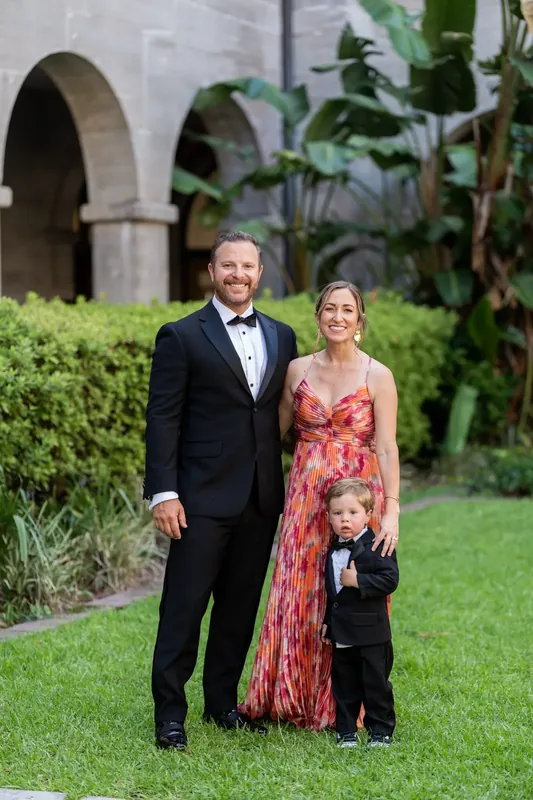 A man in a tuxedo walks across the lawn with a woman in a coral print dress and a young child between them.