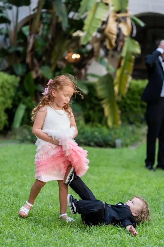 A young flower girl in a pink tutu playfully tugs on a child in a black suit across the courtyard grass.