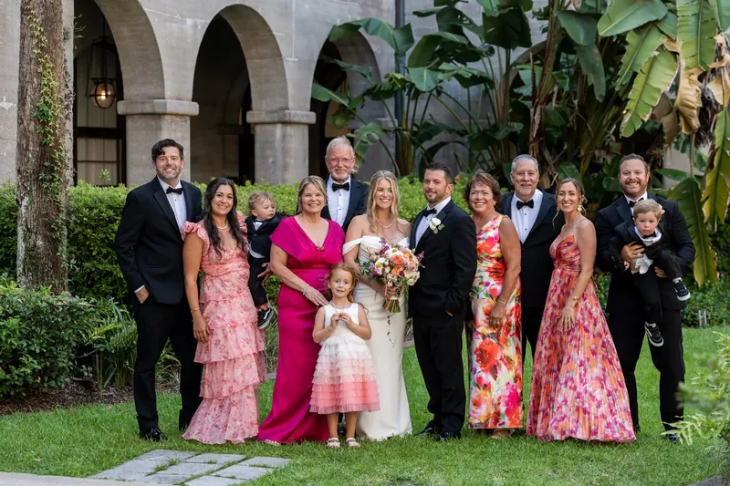 Jenna in a white gown stands center with Russell and extended family in colorful attire beneath the museum's arched