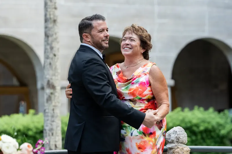 Russell embraces a guest in a colorful floral dress, both smiling, with the museum's arched facade and manicured hedges