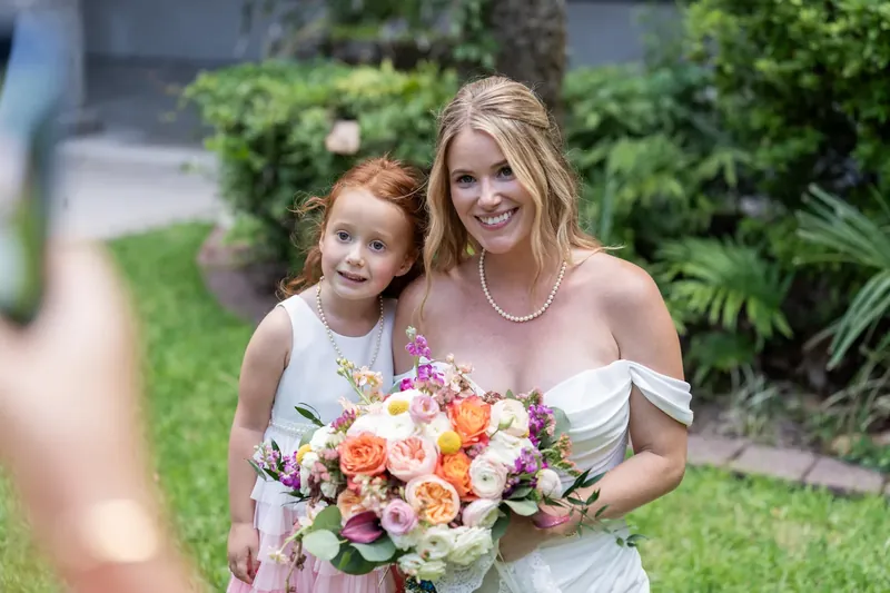 Jenna holds a peach and purple bouquet while posing beside a young girl in a pink dress on the manicured courtyard lawn.