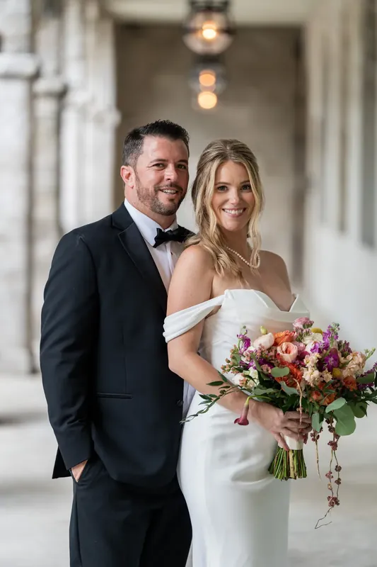 Jenna and Russell smile at the camera while holding her cascading bouquet of coral and blush flowers beneath the museum's