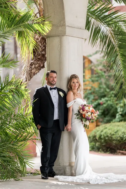 Russell and Jenna pose beneath a tall palm tree and stone column, her bouquet visible as they stand in the courtyard's lush