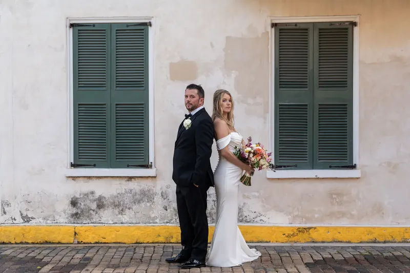 Russell and Jenna pose together against a weathered white building with green shutters and a yellow stripe.