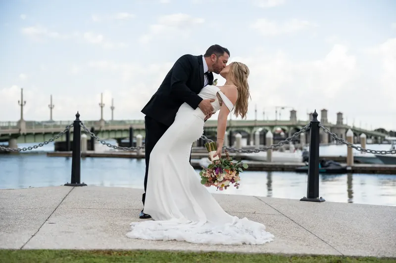 Russell dips Jenna as they kiss on the bayfront promenade, her white gown flowing, moored boats and water stretching behind