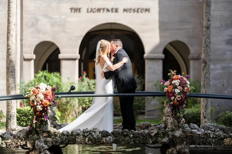 Jenna and Russel embrace under an arched stone opening with burgundy florals on either side.