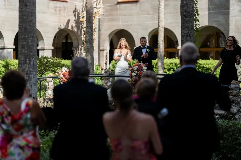 Jenna in a white gown stands before gathered guests with the museum's arches behind her.