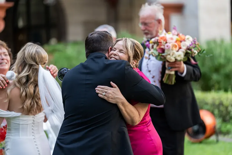 Russel hugs a woman in a hot-pink dress while guests in earth tones embrace nearby on the museum grounds.
