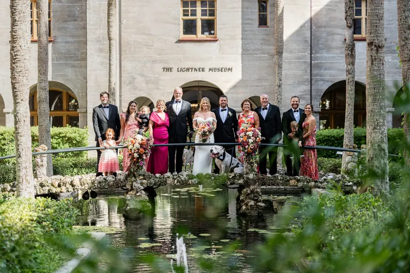 Extended family and wedding party stand together across a reflecting pond in front of the Lightner Museum's stone facade.