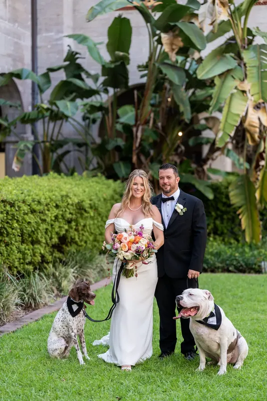 Jenna in a white gown and Russel in a dark suit pose with two dogs holding bouquets at the Lightner Museum Courtyard.