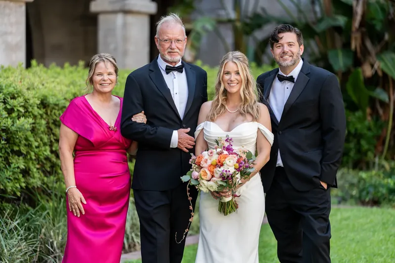 Jenna and Russel stand between two family members in formal attire, holding her colorful bouquet at the Lightner Museum.