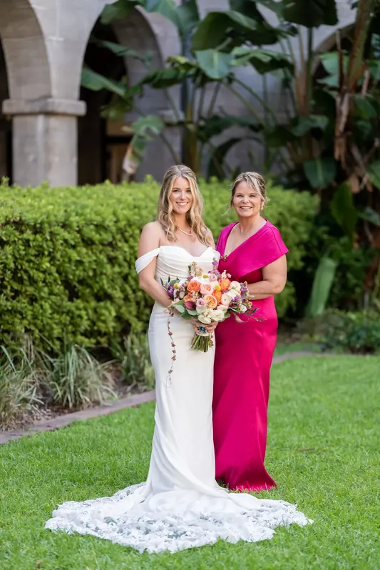 Jenna in white and a partner in magenta hold a coral bouquet at the Lightner Museum Courtyard.