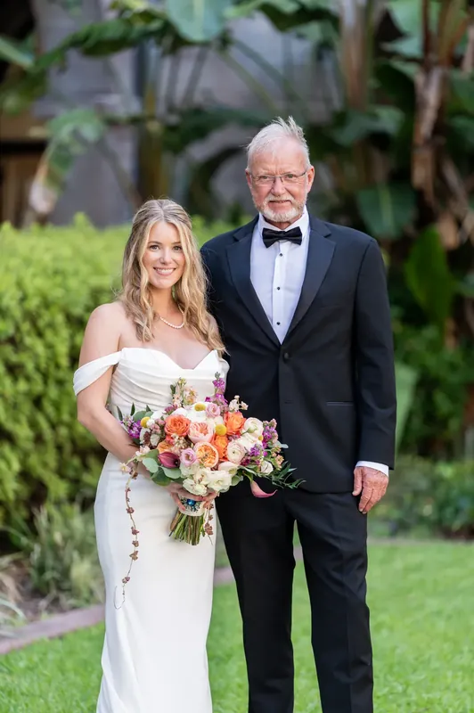 Russell in a tuxedo and Jenna holding their bouquet pose on the lawn surrounded by arched architecture and lush greenery.