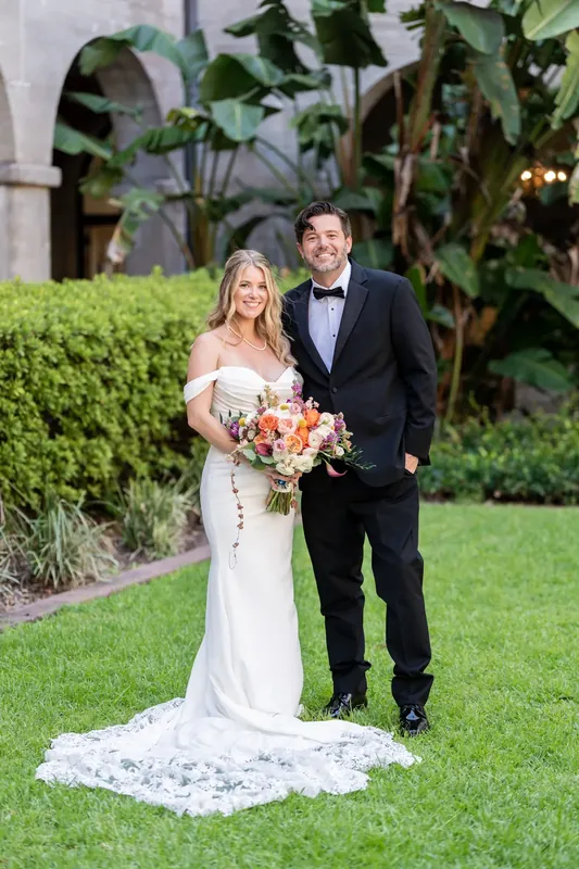 Jenna and Russell stand on the grass holding their bouquet, he in a bow tie and dark suit.