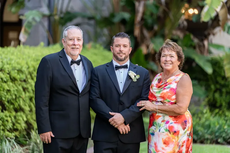 Russell in a tuxedo with boutonniere stands with his father and mother at the Lightner Museum Courtyard.