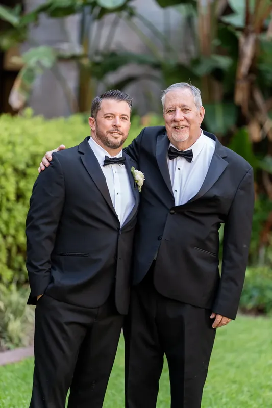 Russell and his father stand side by side in matching black tuxedos with boutonnieres at the ceremony venue.