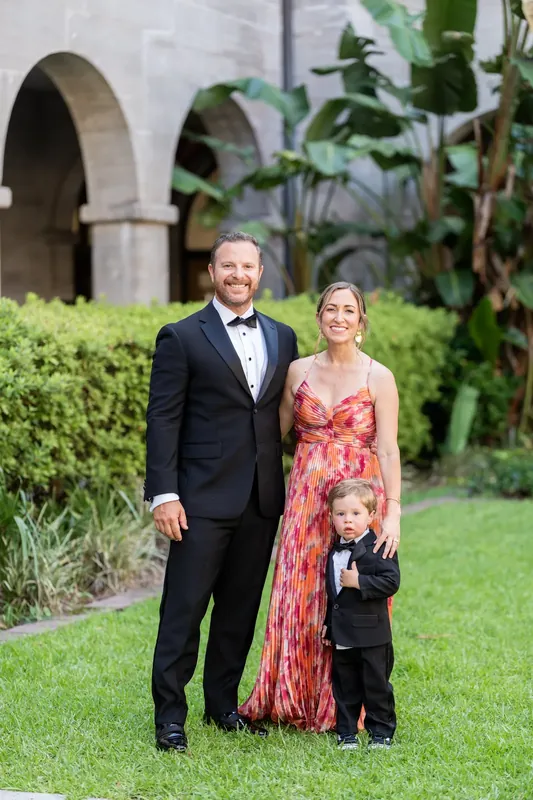 Russell holds hands with Jenna in a coral patterned dress while their young son stands between them on the lawn.