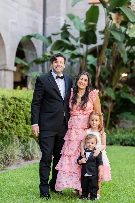 Russell in a black tuxedo stands with Jenna in a pink dress and two children on the lawn at Lightner Museum Courtyard.