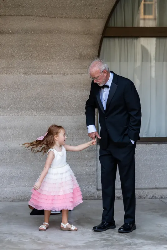 An older man in a tuxedo holds hands with a girl in a white and pink dress on a stone patio.