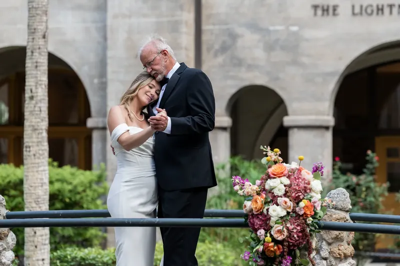 Russell and Jenna embrace near a colorful floral arrangement with "The Lightner" building visible behind them.