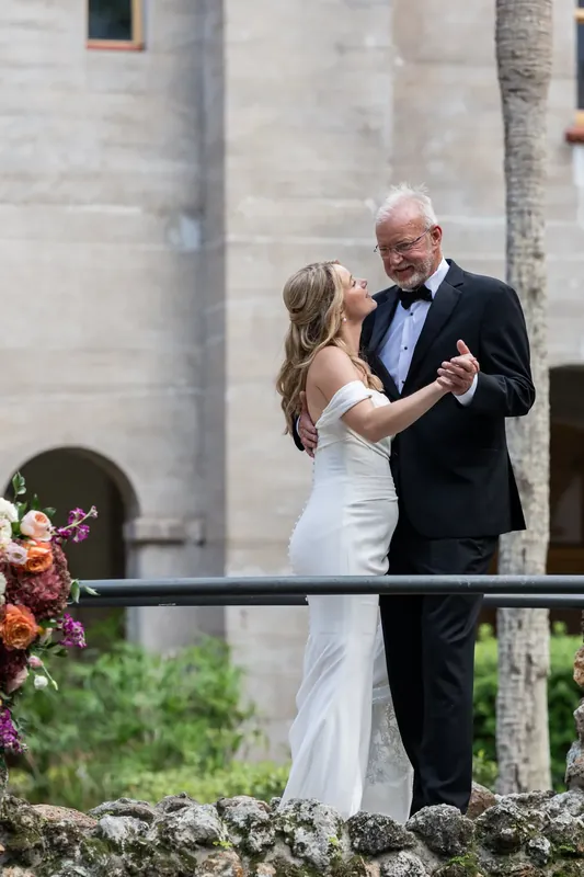 The couple dances together on a stone terrace with vibrant pink and orange flowers beside them.