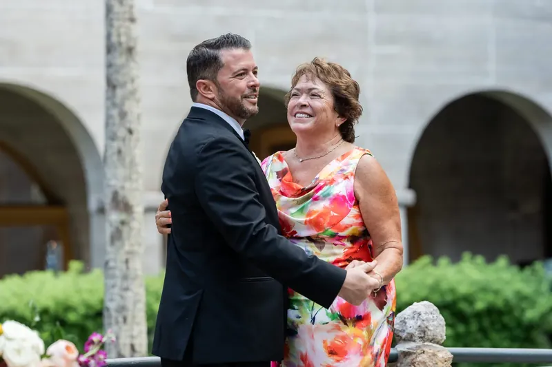 Jenna and Russell smile at each other framed by arched stonework and hedges at the Lightner Museum.
