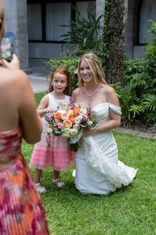 A woman in a white dress kneels on the grass holding a colorful bouquet beside a small child in a pink dress.