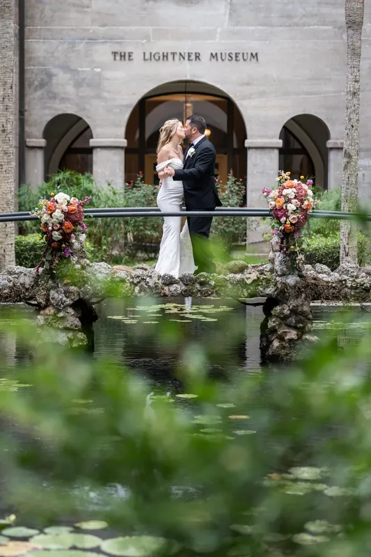 Jenna and Russell kiss over lily-filled water beneath the museum's columned entrance, flanked by floral arrangements.