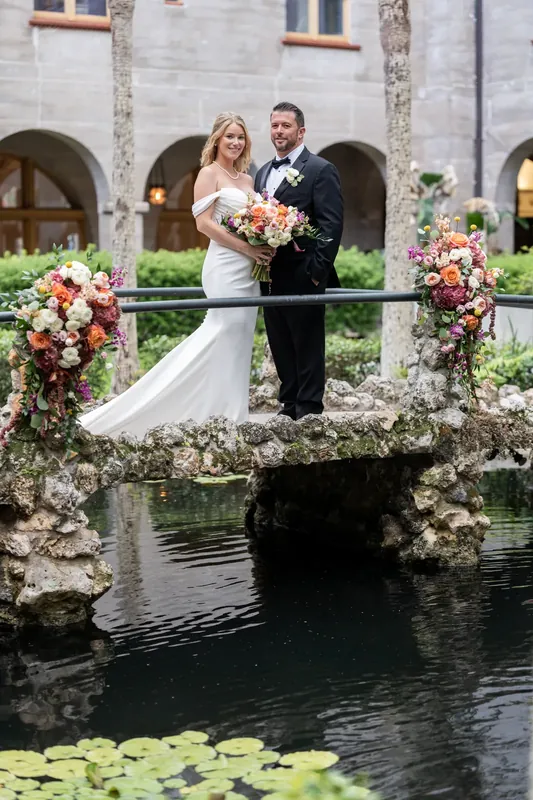 Jenna and Russell stand on a moss-covered bridge with flowering urns and arches behind them.