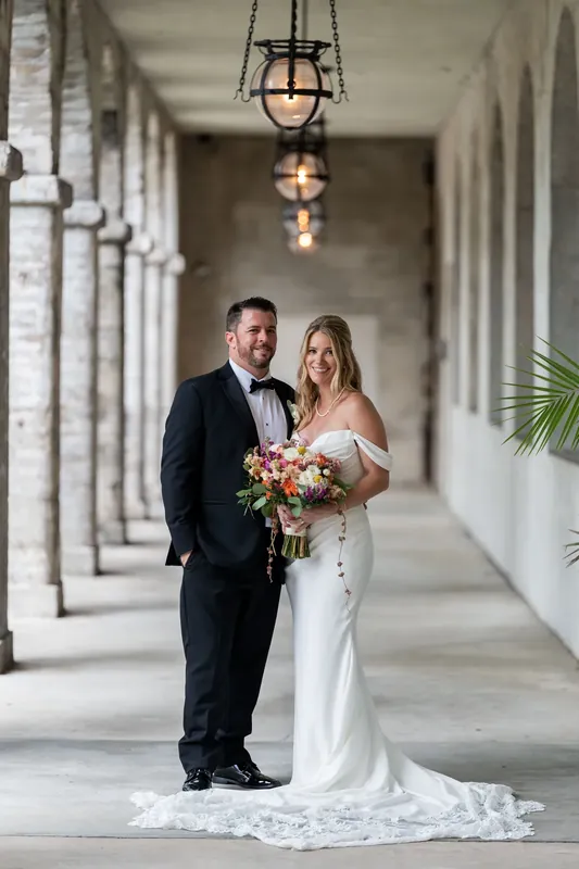 Jenna and Russell stand together in a columned courtyard, she holding a coral and burgundy bouquet while wearing an off-shoulder white gown with a flowing train.