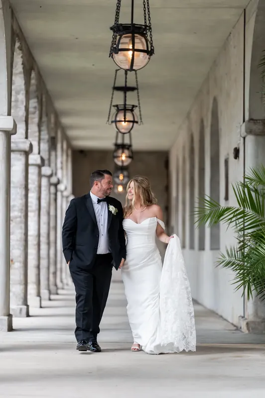 Jenna and Russell walk hand-in-hand down the columned passage, her white dress train trailing across polished stone.