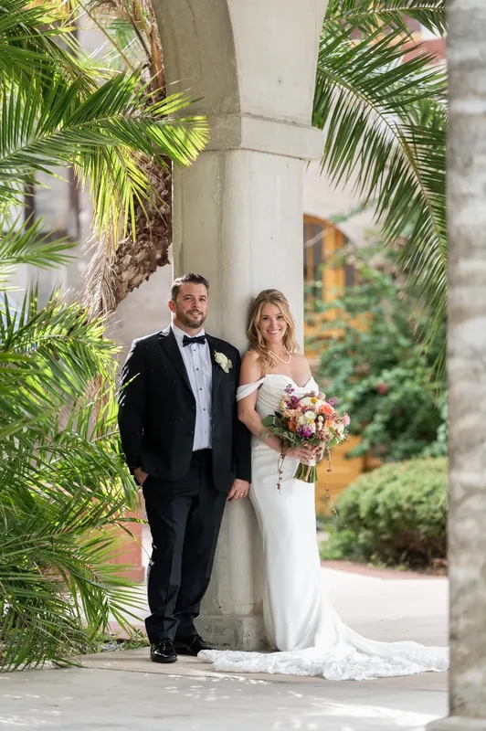 Jenna and Russell stand framed by palm fronds in formal attire against classical columns at the museum courtyard.