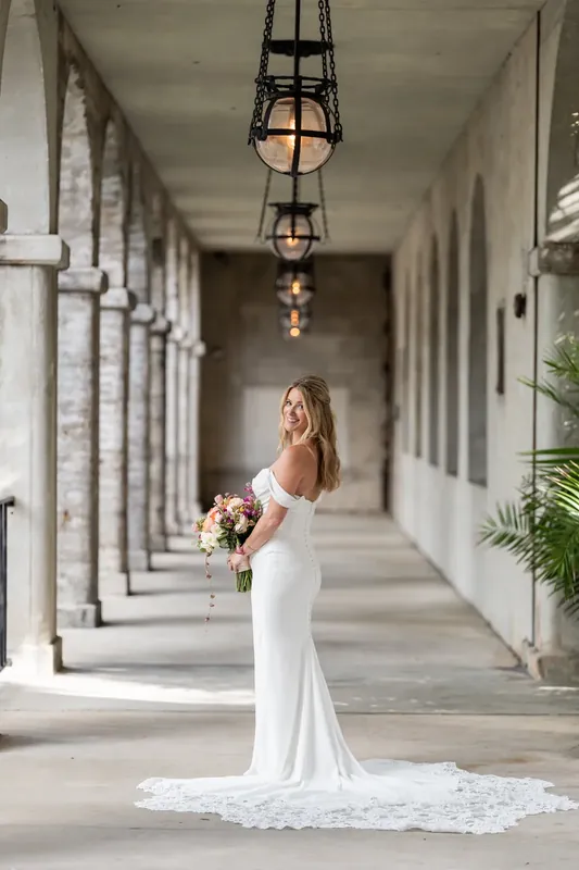 Jenna stands alone in the colonnade in her flowing white gown, holding her bouquet as her train pools behind her.