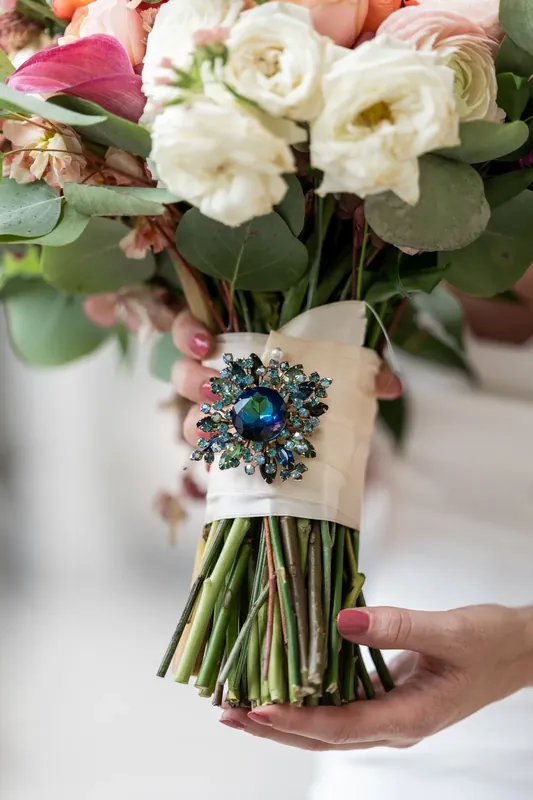 Hands cradle a bridal bouquet featuring cream roses, coral ranunculus, and a blue sapphire brooch.