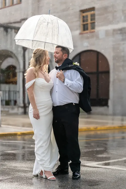 Jenna and Russel huddle under a white umbrella in rain at the Lightner Museum Courtyard.