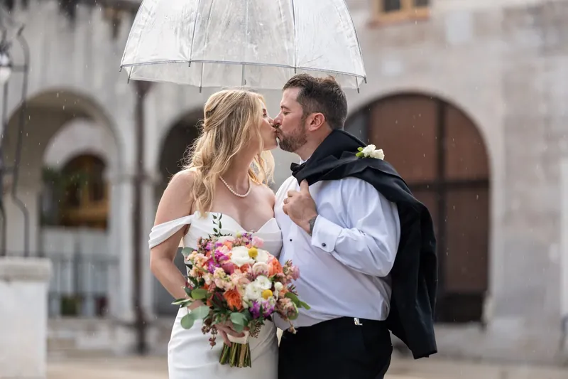 Under their umbrella, Russel gazes at Jenna as she holds a vibrant bouquet, the museum's arched doorways framing the scene.