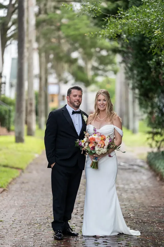 Russel walks beside Jenna down a tree-lined street, she in her white gown holding her bouquet, both smiling at each other.