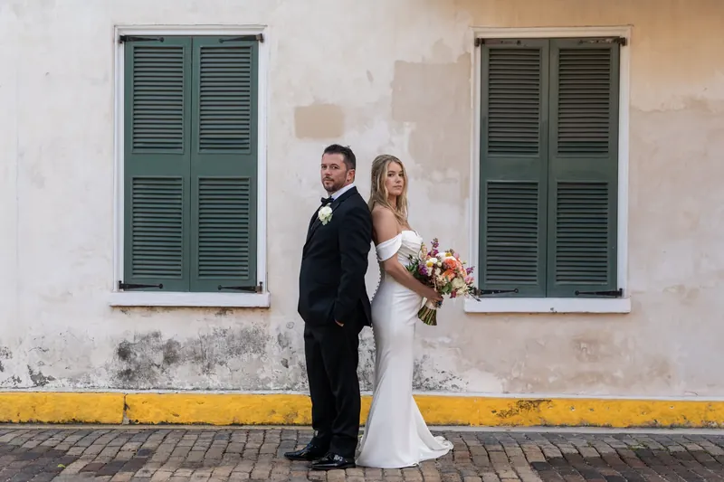 Russel and Jenna stand against a cream wall with green shutters, her bouquet at her side.