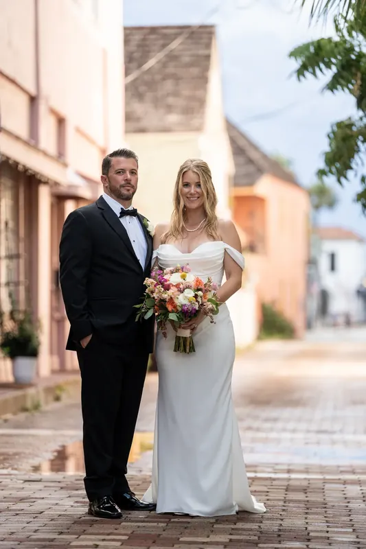 Jenna and Russel pose on a brick plaza with her colorful bouquet and colonial buildings behind them.