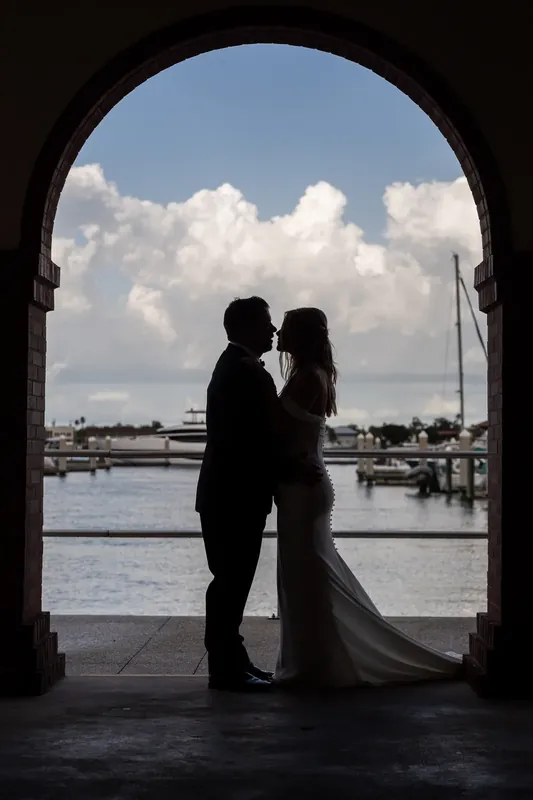 Jenna and Russell kiss in silhouette beneath an archway at St. Augustine Bayfront, with sailboats and water visible beyond.