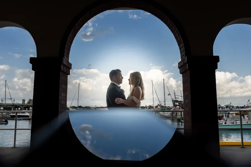 The couple embraces through a circular stone opening framing the marina and moored boats behind them.