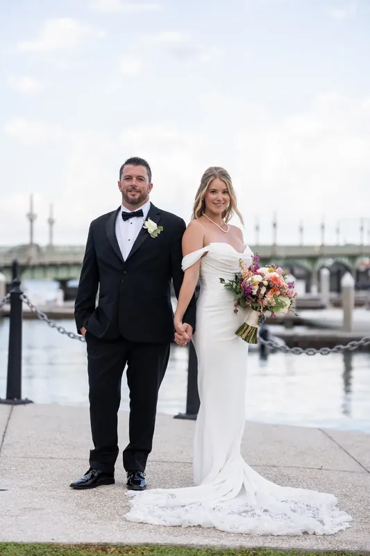Russell in a dark tuxedo and Jenna in a white off-shoulder gown walk hand-in-hand along the bayfront promenade.