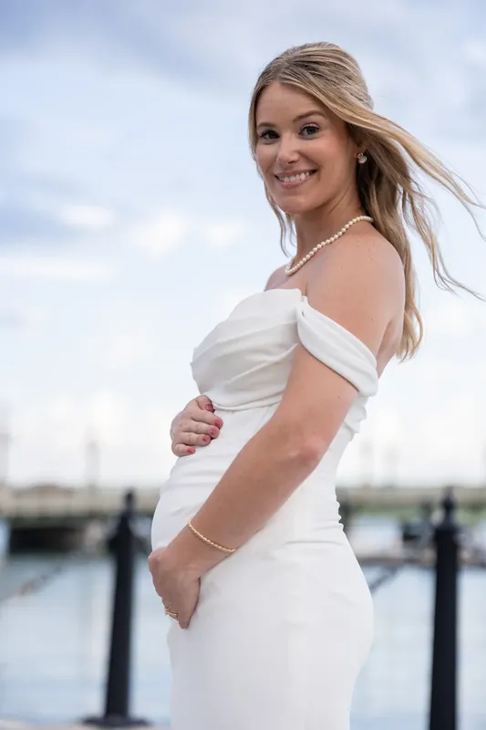 Jenna turns to smile at the camera in her white off-shoulder wedding dress, the bayfront and docked boats behind her.