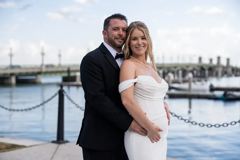 Russell wraps his arm around Jenna from behind as they stand together overlooking the marina at the bayfront.